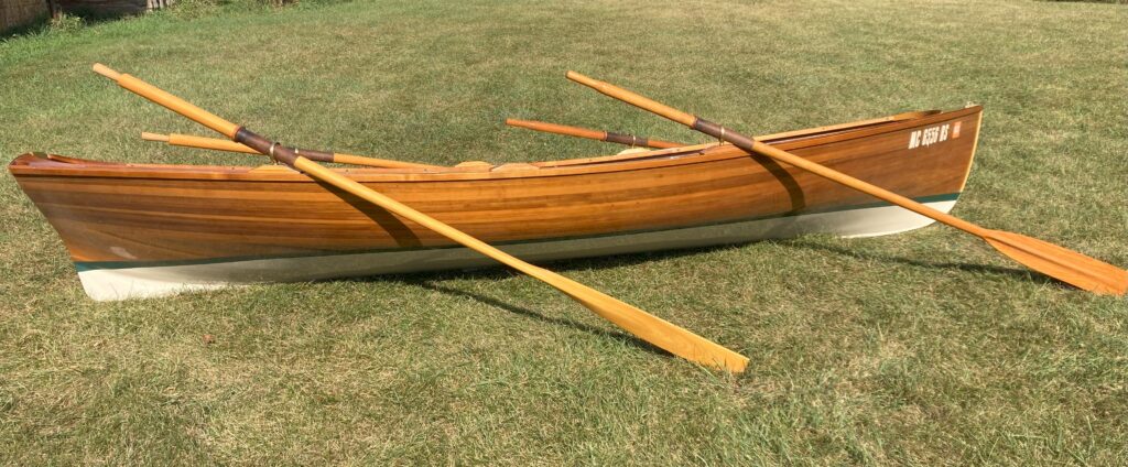 The image shows a wooden rowboat placed on a grass lawn. The boat has a sleek, polished appearance with noticeable wood grain. Two wooden paddles are propped across the middle of the boat.