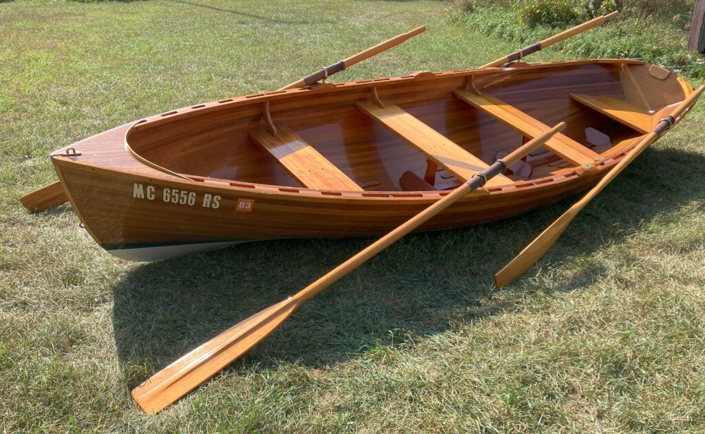 The rowboat in the image is beautifully crafted with a natural, polished wood finish, highlighting the grain patterns. It has two rows of seats across its width, and the interior is spacious with a clean, streamlined design. The oars are made of the same wood tone.